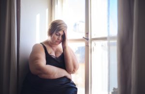 Depressed lonely fat woman in underwear sitting near window,looking down and thinking at home.
